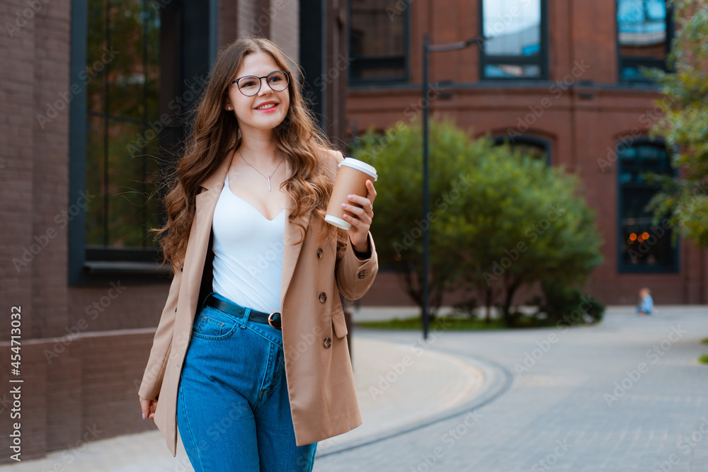 Young charming brunette girl in glasses and stylish blazer walking with cup of coffee in city center Young charming brunette girl in glasses and stylish blazer walking with cup of coffee in city center