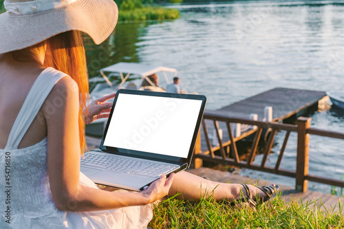 Mock up image of a woman using and typing on a laptop with a blank desktop screen while sitting on the pier.