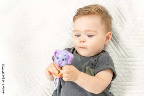 Curious little baby child with rattle toy lying on bed in a sunny bedroom.