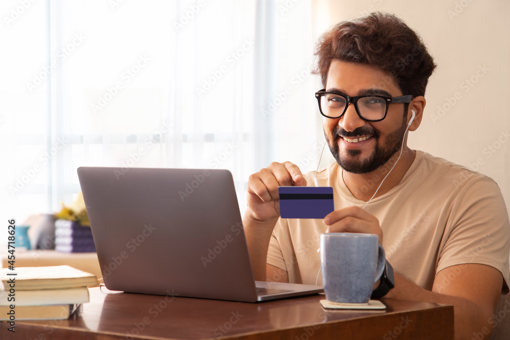 A man with laptop wearing earphones showing credit card and smiling. A man with laptop wearing earphones showing credit card and smiling.
