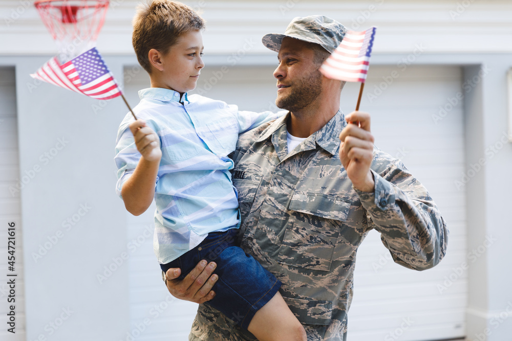 Smiling caucasian male soldier with son outside house holding american flags Smiling caucasian male soldier with son outside house holding american flags