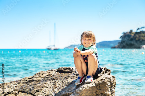 Portrait of a smiling boy looking up, sitting on a rock by the sea in hot weather. Family summer holidays at french seaside. Child wearing sun protection tshirt and swimming shoes.