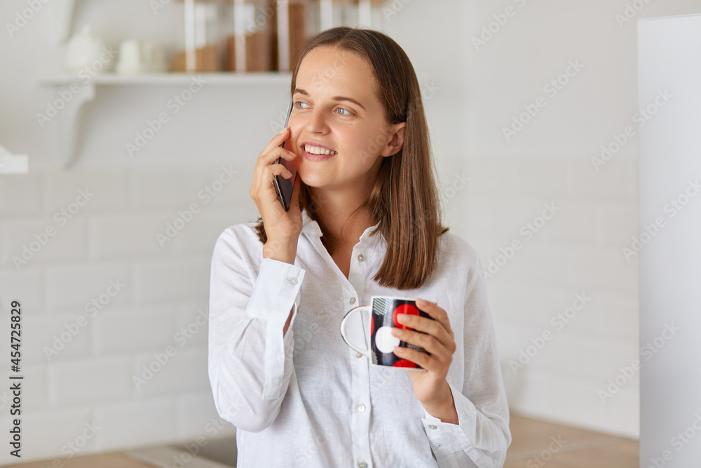 Happy smiling woman talking phone in morning after waking up, drinking hot beverage, enjoying coffee or tea and looking away with optimistic facial expression. Happy smiling woman talking phone in morning after waking up, drinking hot beverage, enjoying coffee or tea and looking away with optimistic facial expression.