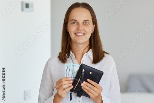 Portrait of beautiful smiling woman in white shirt standing in light room at home, holding wallet with banknotes in hands, looking at camera, wants to buy expensive thing.