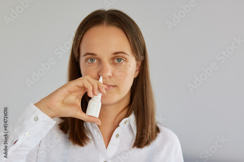 Photo of dissatisfied dark haired woman, uses nasal spray, suffers from stuffy nose, wearing white clothes, posing indoor against light wall, looking at camera.