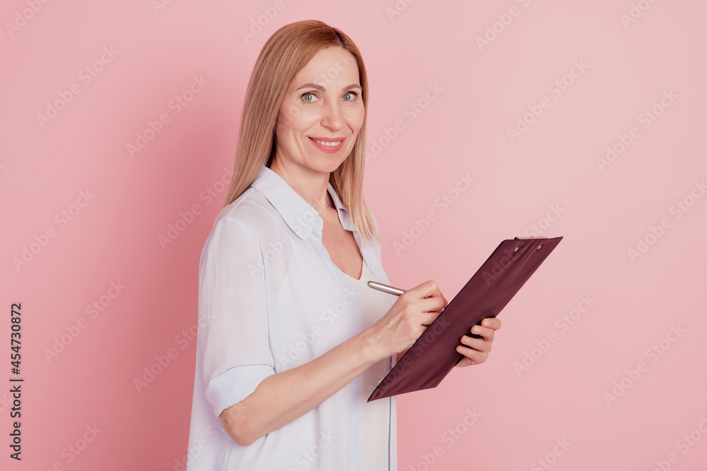 Profile side photo of young business woman happy positive smile hold clipboard write notes isolated over pink color background Profile side photo of young business woman happy positive smile hold clipboard write notes isolated over pink color background