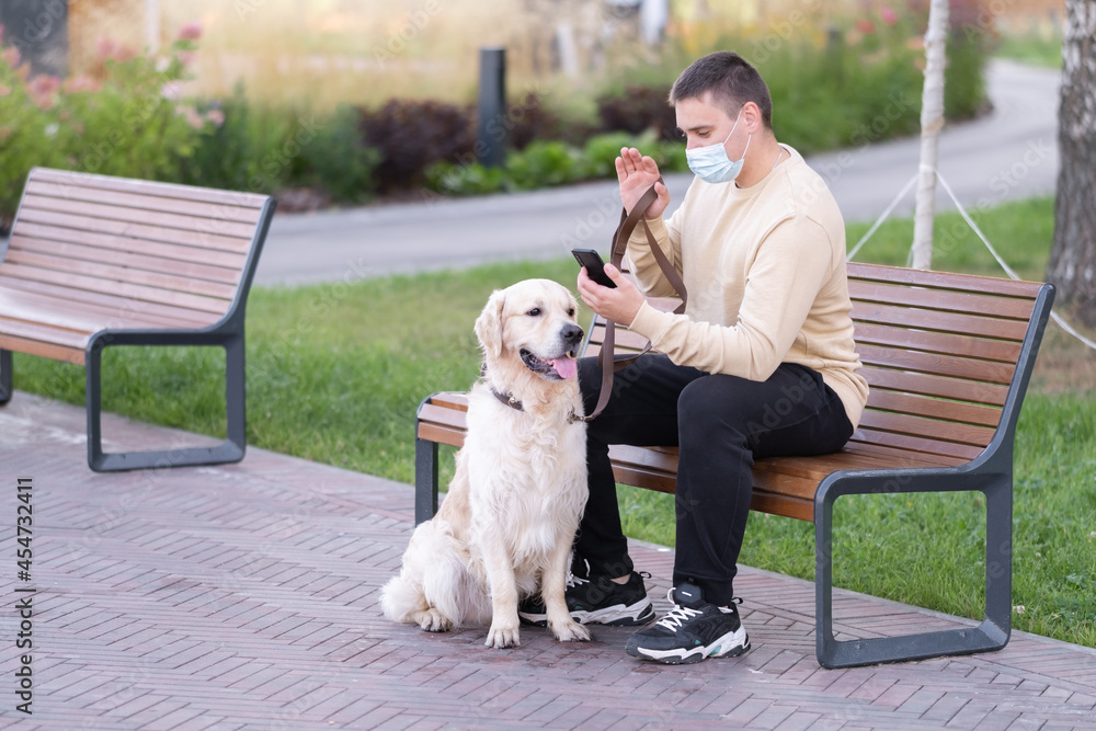 A man sits in a mask from a coronavirus in a park with a dog and looks at the phone. The golden retriever walks with its owner on a sunny day. A man sits in a mask from a coronavirus in a park with a dog and looks at the phone. The golden retriever walks with its owner on a sunny day.