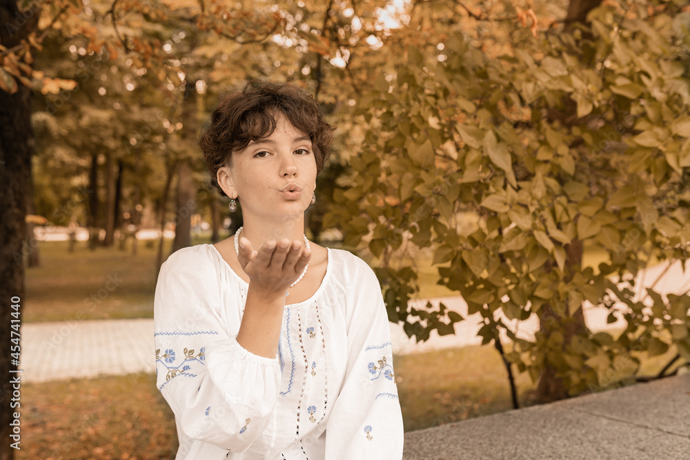 Young attractive slim girl teenager in national embroidered shirt sends an air kiss on blurred background of an autumn park Young attractive slim girl teenager in national embroidered shirt sends an air kiss on blurred background of an autumn park