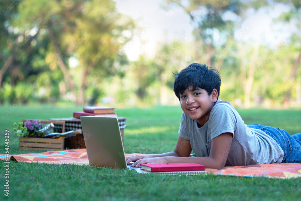 Indian boy lying and using laptop on picnic mat in the garden, outdoor activity concept Indian boy lying and using laptop on picnic mat in the garden, outdoor activity concept