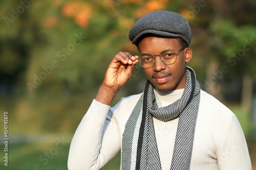 Portrait of handsome intelligent smart guy student, young black Afro African American ethnic man in hat, scarf and glasses walking in beautiful golden autumn park at fall sunny day, looking at camera