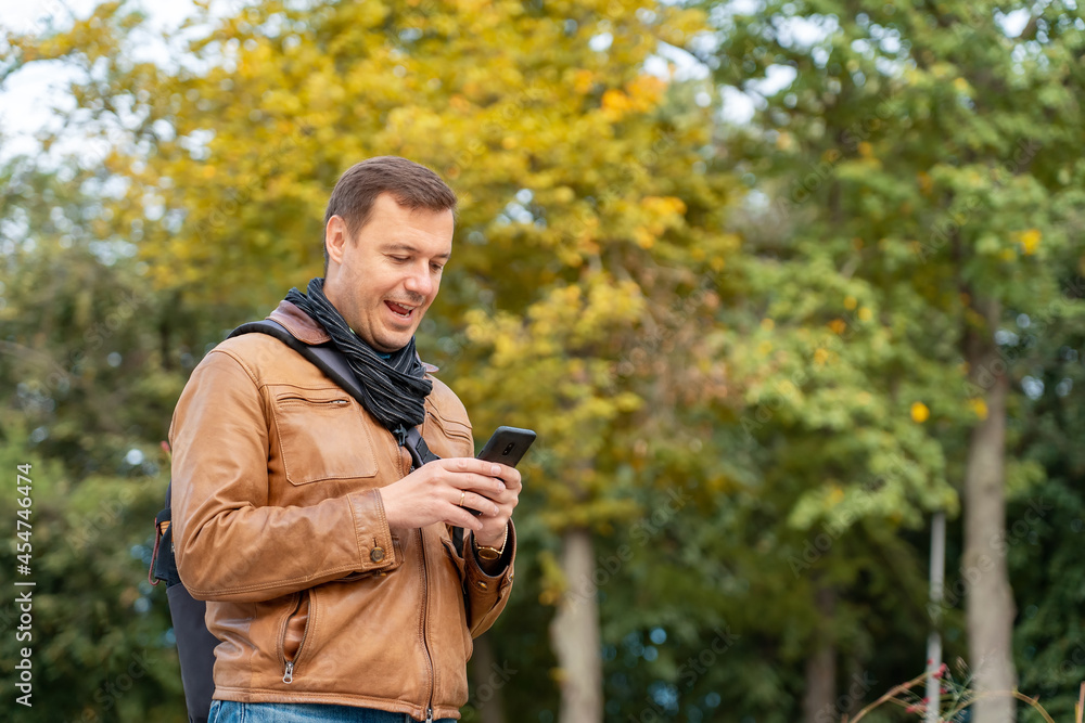 Man with happy face checking phone standing in autumn park outdoors. Portrait of excited man holding smart phone in street. Man with happy face checking phone standing in autumn park outdoors. Portrait of excited man holding smart phone in street.
