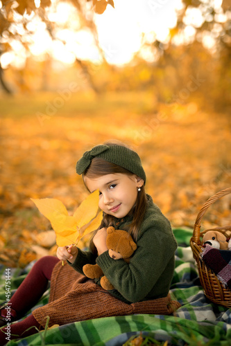 A cute little girl is sitting on a blanket in the park with a toy bear and covers her eye with a dry yellow autumn leaf. A warmfall day in nature. Favorite children s soft plush toys.