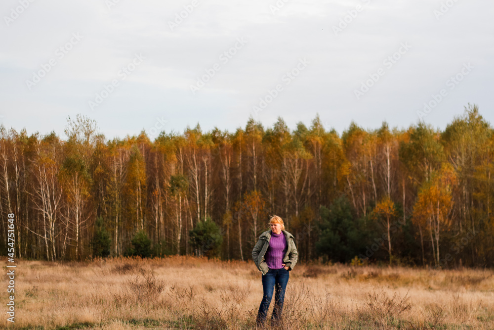 Defocus happy blond 40s woman standing in yellow autumn forest nature background. Happy beautiful lady. Women wearing purple sweater. Fall park, leaves. Dry grass. Out of focus