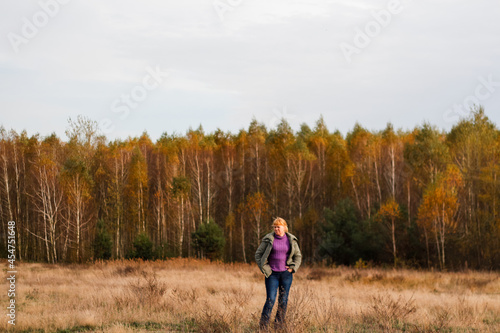 Defocus happy blond 40s woman standing in yellow autumn forest nature background. Happy beautiful lady. Women wearing purple sweater. Fall park, leaves. Dry grass. Out of focus
