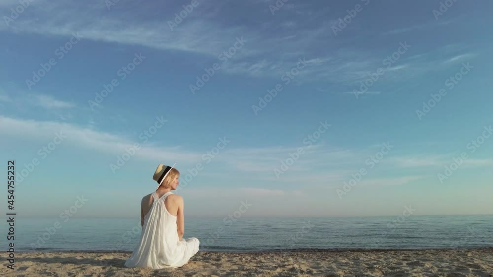Young slim woman wearing white long dress and hat sit alone at beach of calm ocean or sea. Young slim woman wearing white long dress and hat sit alone at beach of calm ocean or sea.
