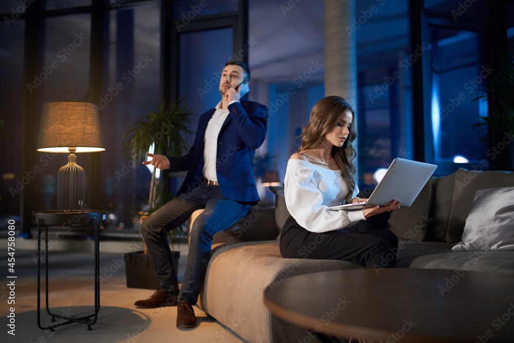 Busy man in stylish suit having mobile conversation while woman in formal clothes working on laptop. Couple solving business issues during evening time at home. Busy man in stylish suit having mobile conversation while woman in formal clothes working on laptop. Couple solving business issues during evening time at home.
