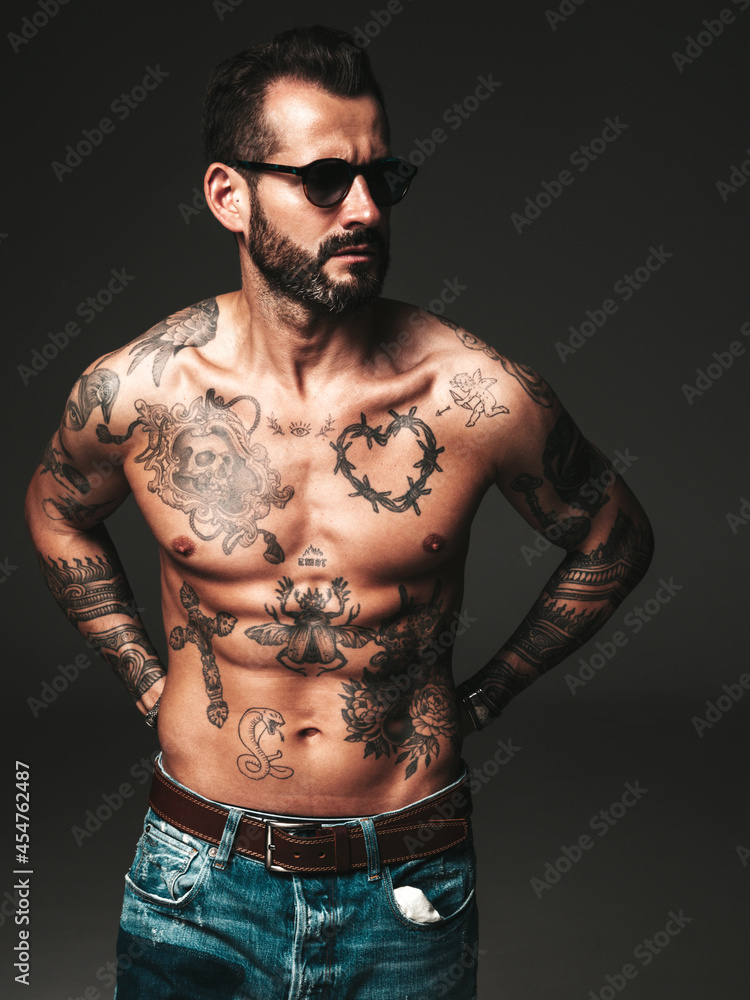 Fototapeta premium Portrait of handsome confident stylish hipster lambersexual model. Sexy modern man. Naked torso with tattoos.Fashion male posing in studio on dark background. In sunglasses