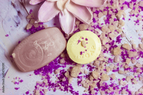 Soap bars and a beautiful lotus flower lay on a white surface, purple lavender-scented sea salt for bath scattered on it flatly. Spa treatments, relax procedures. Body care, beauty concept. Lilac Tone