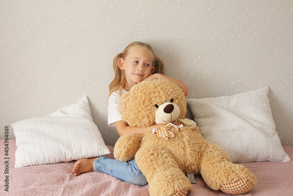 a little blonde girl in a white T-shirt is sitting on the bed and hugging a teddy bear