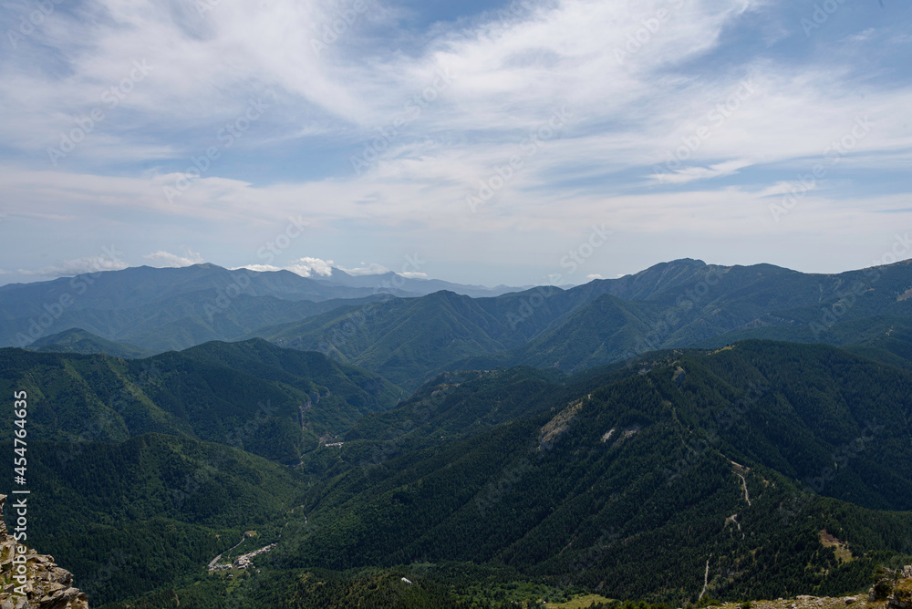 View of the valley from the top of the mountain in front, you can see in the background the peaks of the mountains of the range, the clouds behind the mountains, the blue sky stained with clouds, the View of the valley from the top of the mountain in front, you can see in the background the peaks of the mountains of the range, the clouds behind the mountains, the blue sky stained with clouds, the