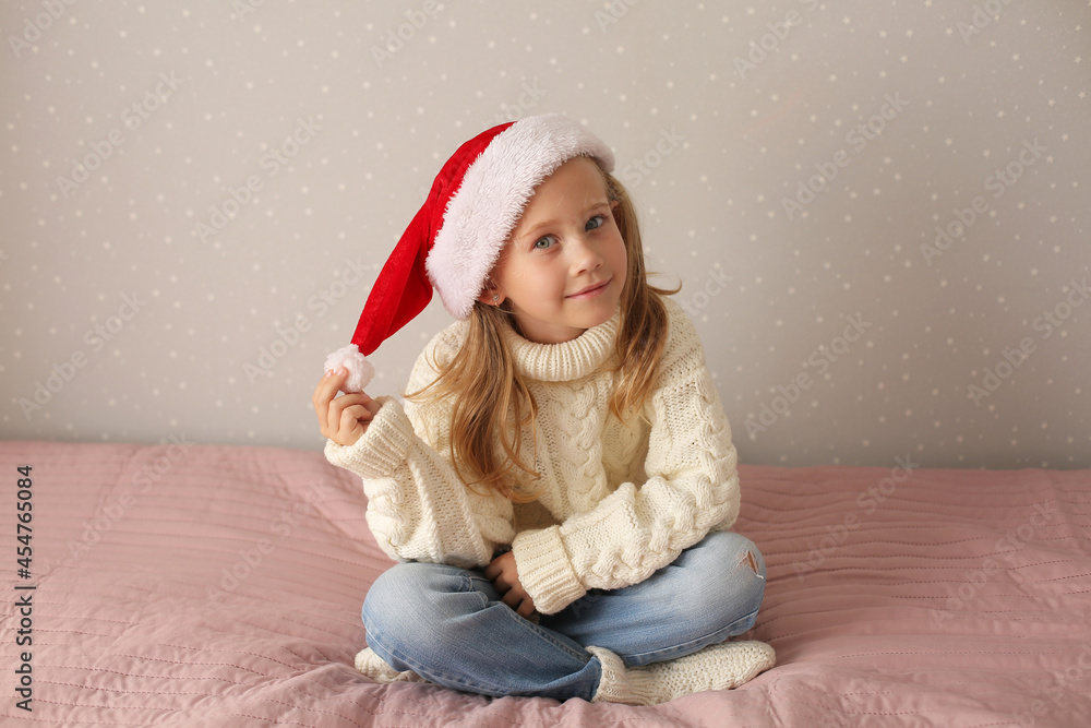 a beautiful little blonde girl is sitting at home on the bed in a winter sweater and a Santa claus hat new year Christmas a beautiful little blonde girl is sitting at home on the bed in a winter sweater and a Santa claus hat new year Christmas