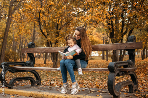 Young female woman babysitter and toddler baby girl read book in autumn park. Happy family mom and toddler outdoors in fall park photo