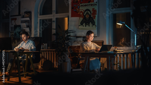 Two Stylish Employees Working on Computers in Creative Agency in Loft Office. Colleagues Answer Emails and Manage Marketing Projects. Dark Renovated Space in Evening with Plants and Artistic Posters.