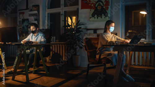 Two Stylish Employees Wearing Protective Face Masks while Working on Computers in Creative Agency in Loft Office. Colleagues Answer Emails and Manage Marketing Projects. Pandemic Covid-19 Concept.