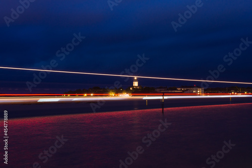 A view towards the sea with an illuminated lighthouse in the background. Light trails of a boat casting reflections on the water.