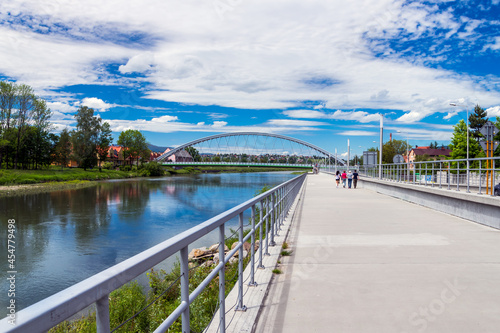 Fototapeta Naklejka Na Ścianę i Meble -  Walking path along the bank of the Koszarawa river. In the background, an arch-shaped bridge. A steel railing separating the sidewalk from the river..Żywiec, Poland
