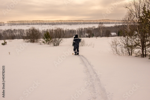 a man on wide skis walks along a hilly area covered with open woodland, lonely houses are visible in the distance