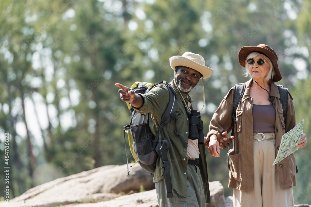 Smiling african american hiker pointing with finger near wife with map in forest.