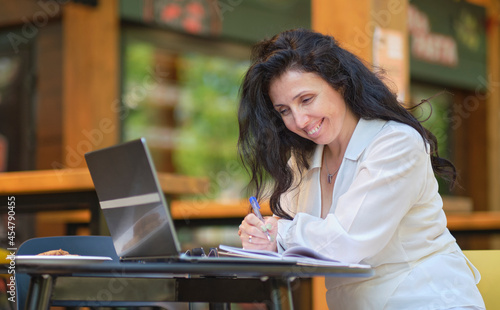 Mature woman at coffee shop. Portrait of confident mature professional woman sitting on summer terrace in cafe, using laptop computer for work, laughing happily indoors. photo