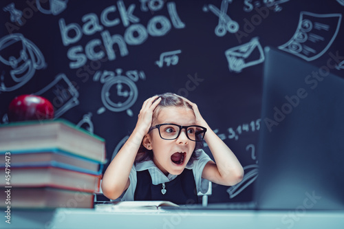 Schoolgirl sits at the table with laptop, books and a chalkboard with school formulas on background. Works homework amazed to put her hands on head, opening mouth wide