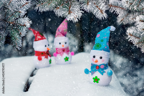 Three toy snowmen in the snow under a spruce tree during a snowfall. New Year and Christmas card