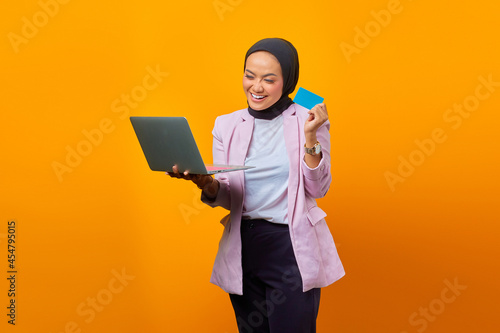 happy asian woman holding laptop and showing blank card over yellow background