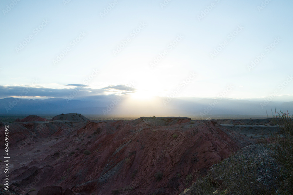 The red canyon and valley at sunset. Panorama view of the arid desert, sandstone formations, rocky mountains, lens flare and hiding sun in Talampaya national park in La Rioja, Argentina.	