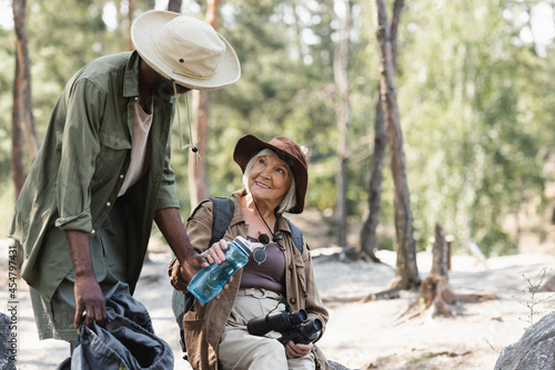 African american hiker holding bottle of water near smiling wife with binoculars in forest. © LIGHTFIELD STUDIOS