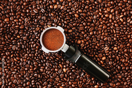  The filter holder for the coffee machine, filled with ground coffee, lies on a background of roasted aromatic coffee beans.