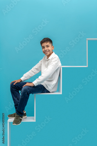 Young smiling teen boy sat down to rest moving up the development or career ladder on a blue background. Education concept