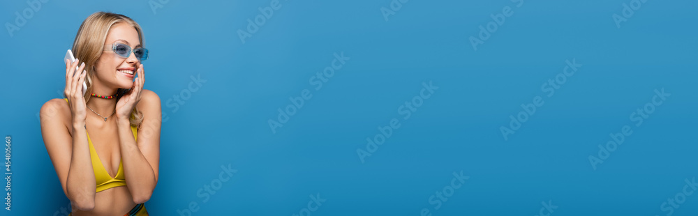 happy young woman in yellow bikini top talking on smartphone isolated on blue, banner. happy young woman in yellow bikini top talking on smartphone isolated on blue, banner.