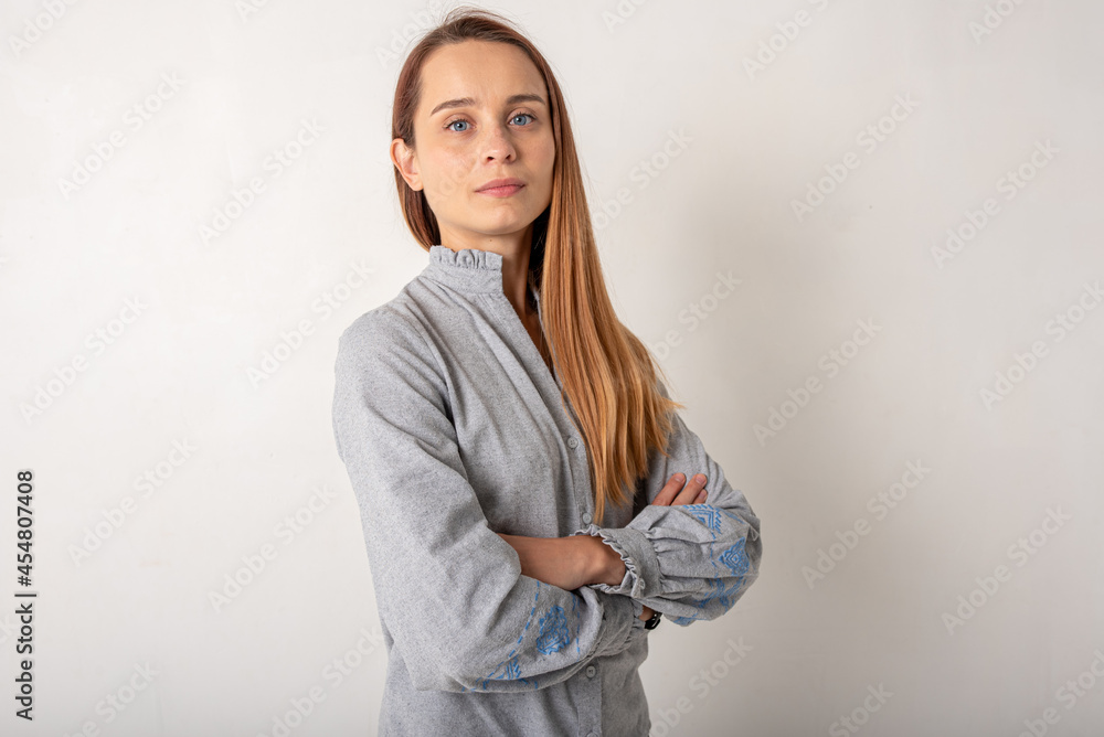 Confident woman in light gray clothes with blue eyes, blond hair and a smile on her face looks at the camera, standing by the white wall with her arms crossed. People, youth, beauty, lifestyle concept Confident woman in light gray clothes with blue eyes, blond hair and a smile on her face looks at the camera, standing by the white wall with her arms crossed. People, youth, beauty, lifestyle concept