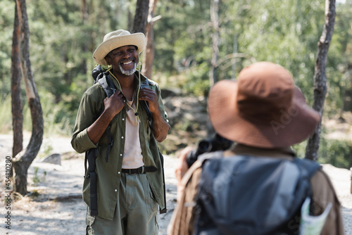 Positive african american traveler looking at blurred wife in hat in forest.