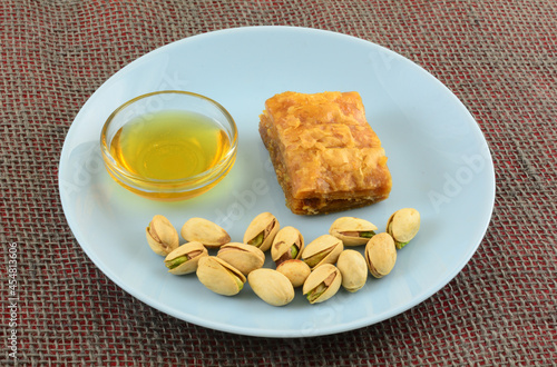 Piece of baklava with honey in glass bowl and pistachio nuts in shells on blue snack plate photo