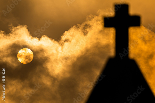 Cemetery cross out of focus in the foreground with clouds and full moon in the background creating a spooky atmosphere, ideal for Halloween.