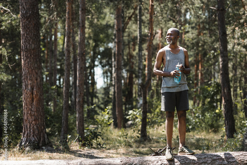 Positive afrian american sportsman holding sports bottle in forest.