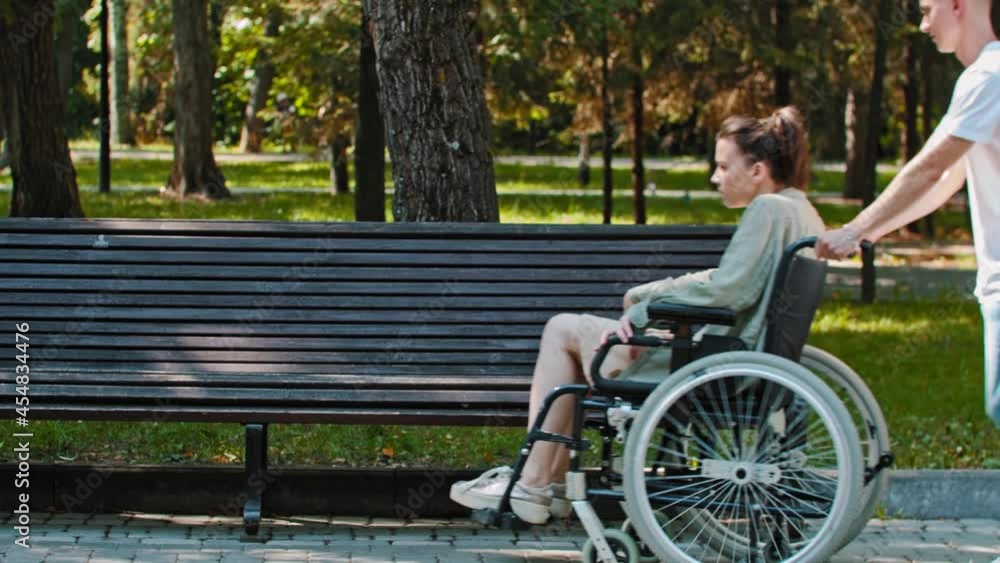 A couple walking in park - a guy rides a woman in a wheelchair and about to transfer her on a bench A couple walking in park - a guy rides a woman in a wheelchair and about to transfer her on a bench