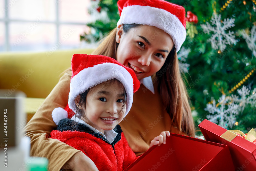 Happy Asian family daughter girl wears sweater red and white Santa Claus hat sitting with mom surprising exciting when open present gift box celebrate Xmas eve near Christmas pine tree in living room Happy Asian family daughter girl wears sweater red and white Santa Claus hat sitting with mom surprising exciting when open present gift box celebrate Xmas eve near Christmas pine tree in living room
