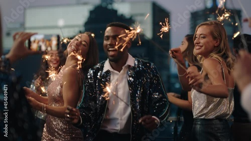 happy fashion friends dancing with sparklers celebrating new years eve at glamorous rooftop party friend using smartphone sharing video of celebration on social media 4k photo