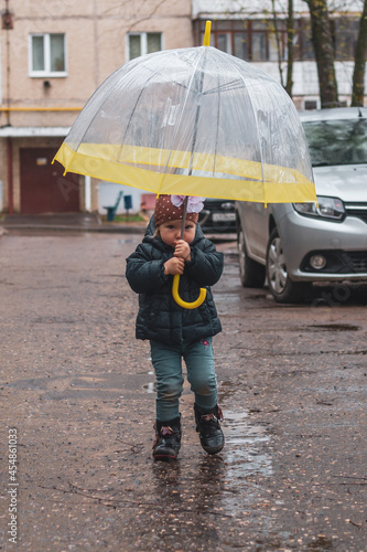 A cute little girl in a blue jacket walks in the cold autumn or spring under a yellow umbrella in rainy weather on the street of the town